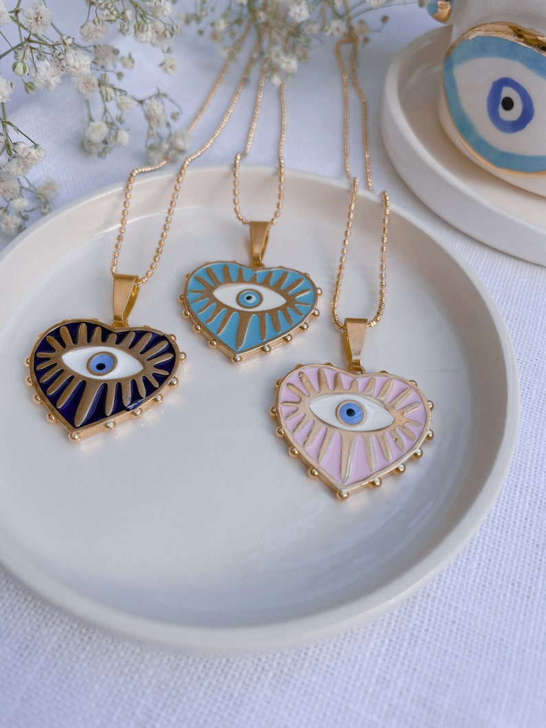 May include: Three gold necklaces with heart-shaped pendants featuring an evil eye design. The pendants are blue, pink, and black with gold accents.