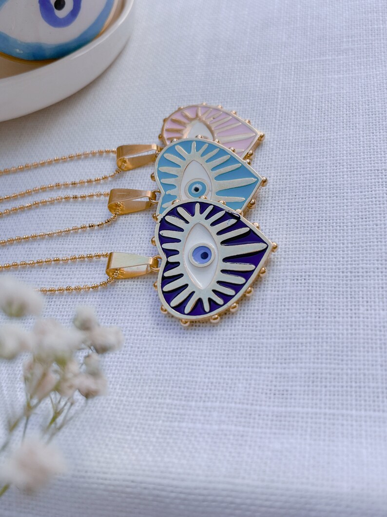 May include: Three gold necklaces with heart-shaped pendants featuring blue evil eyes. The pendants are decorated with white and purple enamel and have a beaded border.