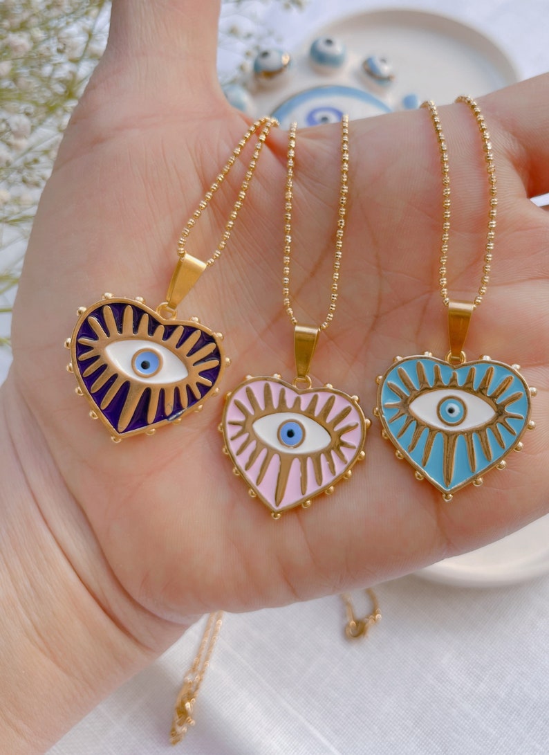 May include: Three gold necklaces with heart-shaped pendants featuring an evil eye design. The pendants are in shades of purple, pink, and blue.