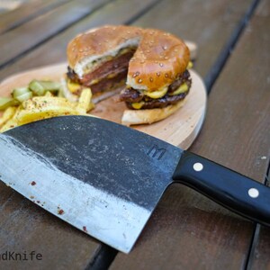 May include: A large, black-handled cleaver with a silver blade sits on a wooden table next to a wooden cutting board with a hamburger, french fries, and pickles on it.