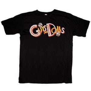 May include: Black t-shirt with the words "Guys and Dolls" in a retro font. The text is in white, red, and yellow with dice graphics. The shirt is a classic crew neck style.