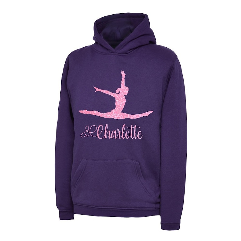 Puede incluir: Sudadera con capucha morada con una silueta de gimnasta de purpurina rosa y el nombre "Charlotte" en letras de purpurina rosa.