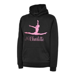 Puede incluir: Sudadera con capucha negra con un dise&ntilde;o de gimnasta haciendo una divisi&oacute;n en purpurina rosa. El nombre "Charlotte" est&aacute; escrito en purpurina rosa debajo de la gimnasta.