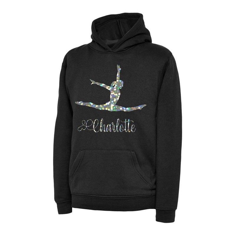 Puede incluir: Sudadera con capucha negra con una silueta de gimnasta de purpurina plateada y el nombre "Charlotte" en letra cursiva.