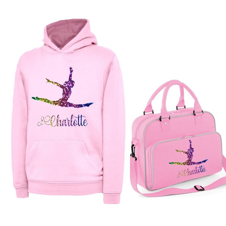 Puede incluir: Sudadera con capucha rosa con una silueta de gimnasta arco&iacute;ris brillante y el nombre "Charlotte" impreso en la parte delantera. Tambi&eacute;n se muestra una bolsa de deporte rosa a juego con el mismo dise&ntilde;o.