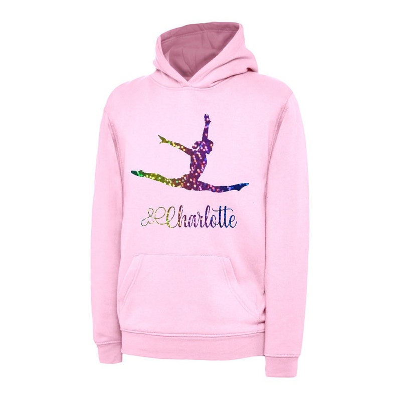 Puede incluir: Sudadera con capucha rosa con una silueta de gimnasta de purpurina arco&iacute;ris y el nombre "Charlotte" en una fuente cursiva.