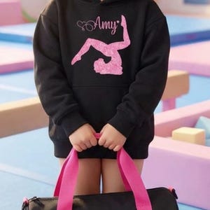 Könnte beinhalten: Ein junges Mädchen trägt einen schwarzen Kapuzenpullover mit dem Namen "Amy" und einem rosa Gymnastik-Motiv. Sie hält eine schwarze Sporttasche mit rosa Trägern, ebenfalls mit "Amy" und einem Gymnastik-Design. Der Hintergrund zeigt Turngeräte.