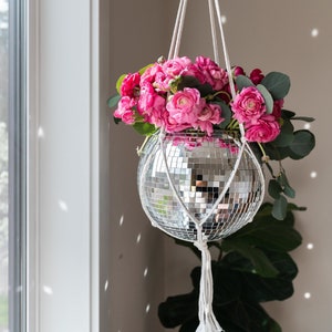 May include: A pink flower arrangement hangs from a white macrame plant hanger. The planter is a silver disco ball.