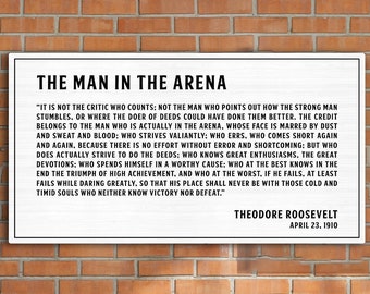 May include: A framed print with the text "THE MAN IN THE ARENA" by Theodore Roosevelt. The quote is in black font on a white background. The print hangs above a black leather sofa in a room with a brick wall.