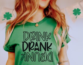 May include: A green t-shirt with the text "DRINK DRANK DUNK" in black. The shirt is being worn by a person sitting on a red stool. The person is wearing black jeans and white sneakers.
