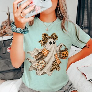 May include: A light mint green t-shirt featuring a central graphic of a ghost adorned with a leopard print bow. The ghost is decorated with various patches including plaid, floral, and leopard patterns, and is holding a small pumpkin bucket.