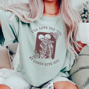 Puede incluir: Una camiseta azul claro con un gr&aacute;fico de dos esqueletos abraz&aacute;ndose. El texto en la camiseta dice "I'll love you 'til my lungs give out."