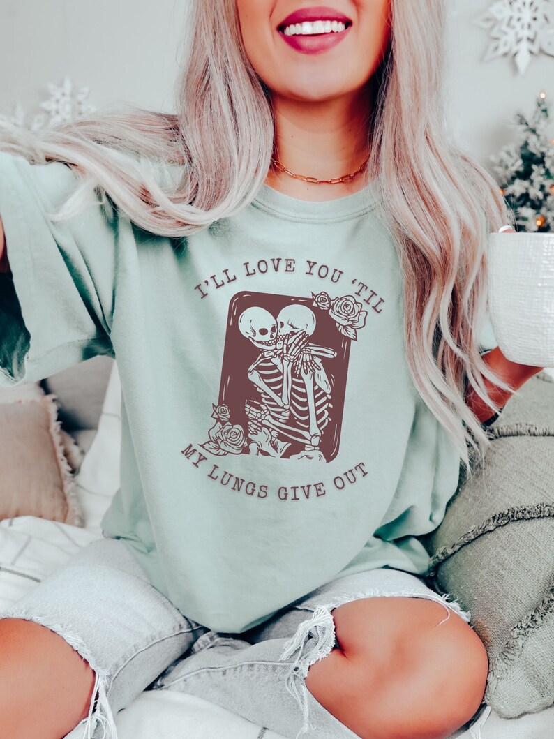 Puede incluir: Una camiseta azul claro con un gr&aacute;fico de dos esqueletos abraz&aacute;ndose. El texto en la camiseta dice "I'll love you 'til my lungs give out."