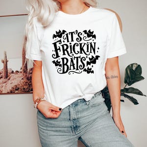 May include: A white short-sleeved t-shirt featuring black graphic text that reads IT'S FRICKIN BATS, surrounded by small illustrations of flying bats and decorative stars. The shirt has a crew neckline and is worn with light blue denim jeans.