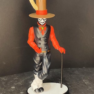 Puede incluir: Una figura negra y roja con una máscara blanca, que lleva un sombrero de copa marrón con una pluma, sosteniendo un bastón negro. La figura está de pie sobre una base blanca con el texto "My Hero Academia" en japonés.