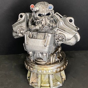 May include: A detailed, silver-colored bust of a power armor helmet from the Fallout video game series. The helmet features multiple lenses, a breathing apparatus, and intricate mechanical details. The base has a weathered, metallic appearance.