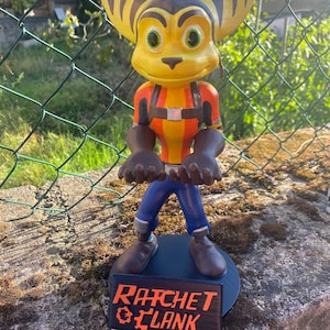 May include: A 3D-printed Ratchet figure from the Ratchet & Clank game. The figure is predominantly yellow, orange, and blue, with large ears and green eyes. The base features the game's logo in orange text on a black background.