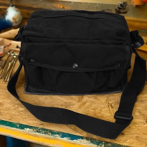 May include: A black canvas messenger bag with a large flap and a smaller pocket on the front. The bag has a long adjustable strap and a buckle closure.