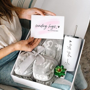 May include: A white gift box filled with a gray blanket with the words "You got this" printed on it, a white tumbler with the words "You got this" printed on it, a small potted succulent plant, and a pair of blue socks. A card with the words "sending hugs... in a little box" is visible in the background.