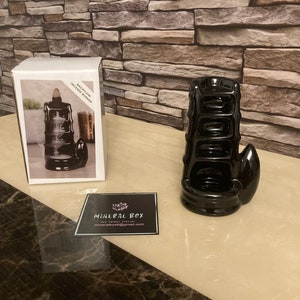 Op de afbeelding: Een zwarte backflow wierookbrander met een uniek gelaagd ontwerp en een gebogen handvat. De brander wordt weergegeven naast de verpakking, met een foto van het product en de woorden "BACKFLOW INCENSE BURNER". Er is ook een visitekaartje aanwezig.