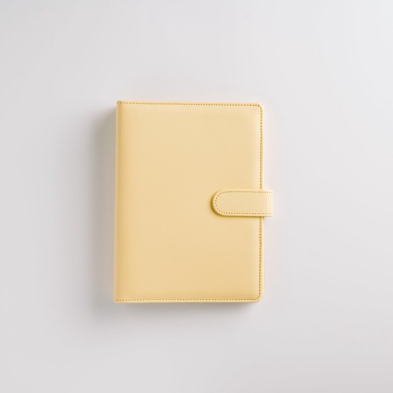 May include: A pale yellow, rectangular planner with a matching tab closure. The planner has a smooth, leather-like texture and is stitched around the edges. The tab closure is secured with a hidden snap.