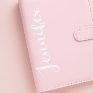 May include: A light pink binder with the name "Jennifer" in white script. The binder has a snap closure and is made of a smooth material. The background is a matching light pink.