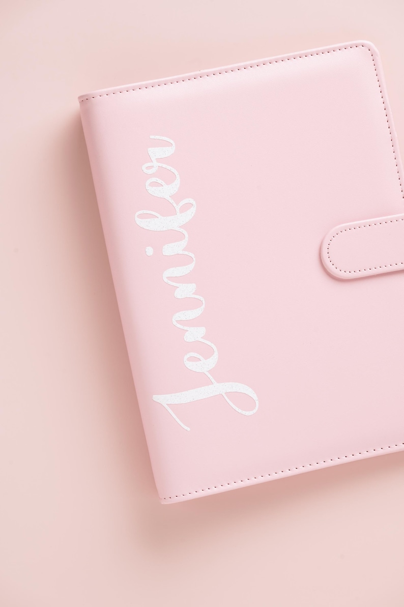May include: A light pink binder with the name "Jennifer" in white script. The binder has a snap closure and is made of a smooth material. The background is a matching light pink.