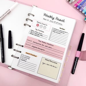 May include: A pink planner with a weekly reset page, featuring sections for mental check-in, top priorities, intentions, and a checklist. The page includes handwritten notes and the text "Weekly Reset". A black pen and highlighter are visible.