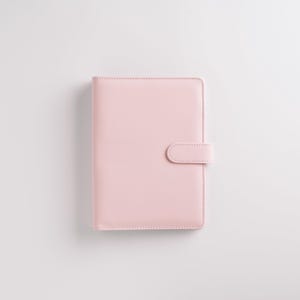 May include: A light pink planner with a smooth, leather-like texture. The cover features a simple, minimalist design with a rounded tab closure on the right side. The planner is closed and appears to be new.