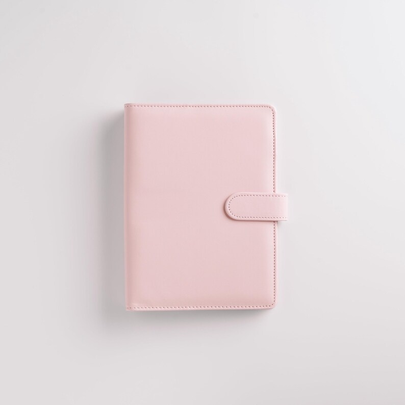 May include: A light pink planner with a smooth, leather-like texture. The cover features a simple, minimalist design with a rounded tab closure on the right side. The planner is closed and appears to be new.