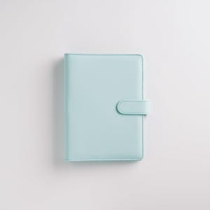 May include: A light blue planner with a matching tab closure. The cover is smooth and the edges are neatly stitched. The planner is rectangular and sits on a white background.