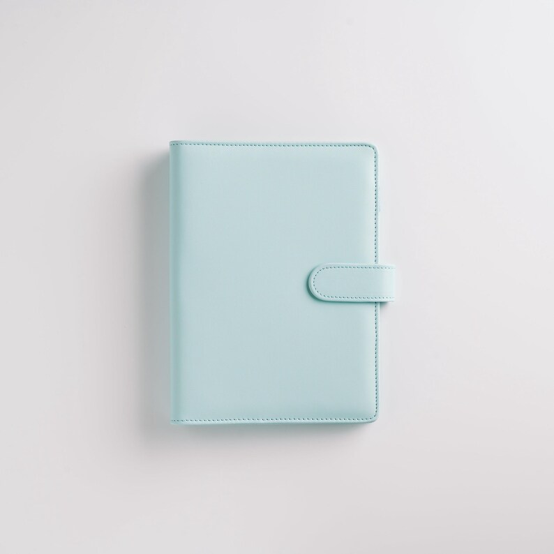 May include: A light blue planner with a matching tab closure. The cover is smooth and the edges are neatly stitched. The planner is rectangular and sits on a white background.