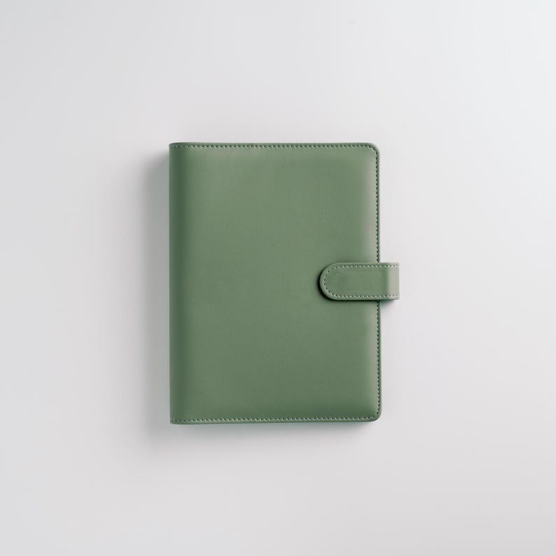 May include: A sage green leather-bound planner with a matching clasp. The cover is smooth and rectangular, with a rounded clasp closure on the right side. The planner is set against a white background.