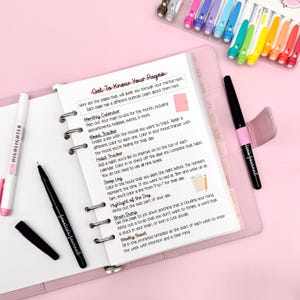 May include: A pink planner with a handwritten guide titled "Get To Know Your Pages." The open pages display instructions for monthly calendars, mood trackers, and habit trackers. A black pen and a highlighter are visible.