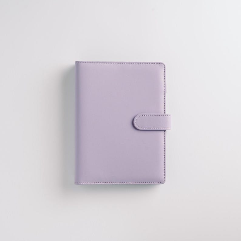 May include: A lavender-colored planner with a snap closure. The cover is smooth and the edges are neatly stitched. The planner is rectangular and appears to be closed. The background is plain white.