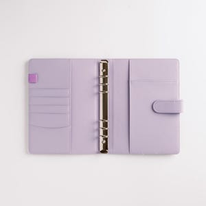 May include: Open, lavender-colored binder with a silver metal ring mechanism. The interior features card slots, a pocket, and a tab closure. A small purple elastic band is on the left side. The binder is on a white background.