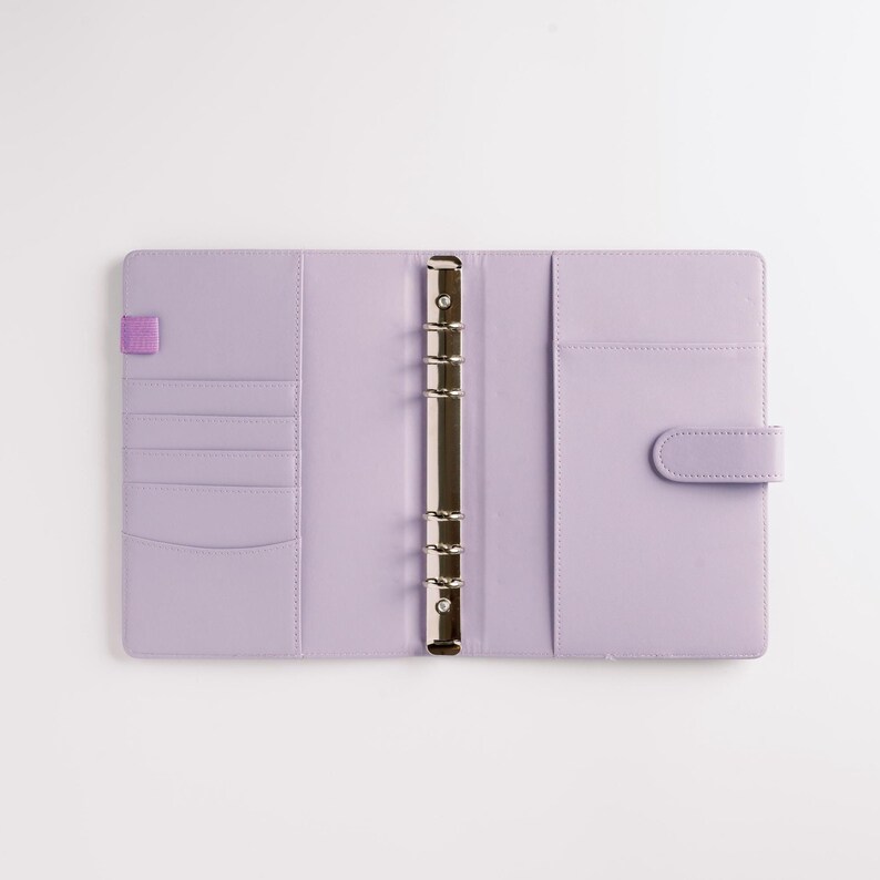 May include: Open, lavender-colored binder with a silver metal ring mechanism. The interior features card slots, a pocket, and a tab closure. A small purple elastic band is on the left side. The binder is on a white background.