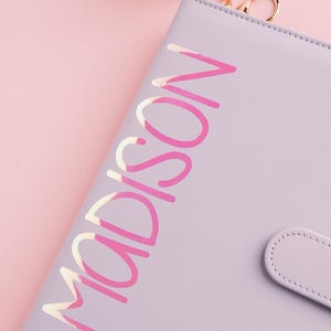 May include: A lavender-colored planner with the name "MADISON" in a pink and white script. The planner has a matching snap closure and a fluffy pink keychain. The background is a soft pink.