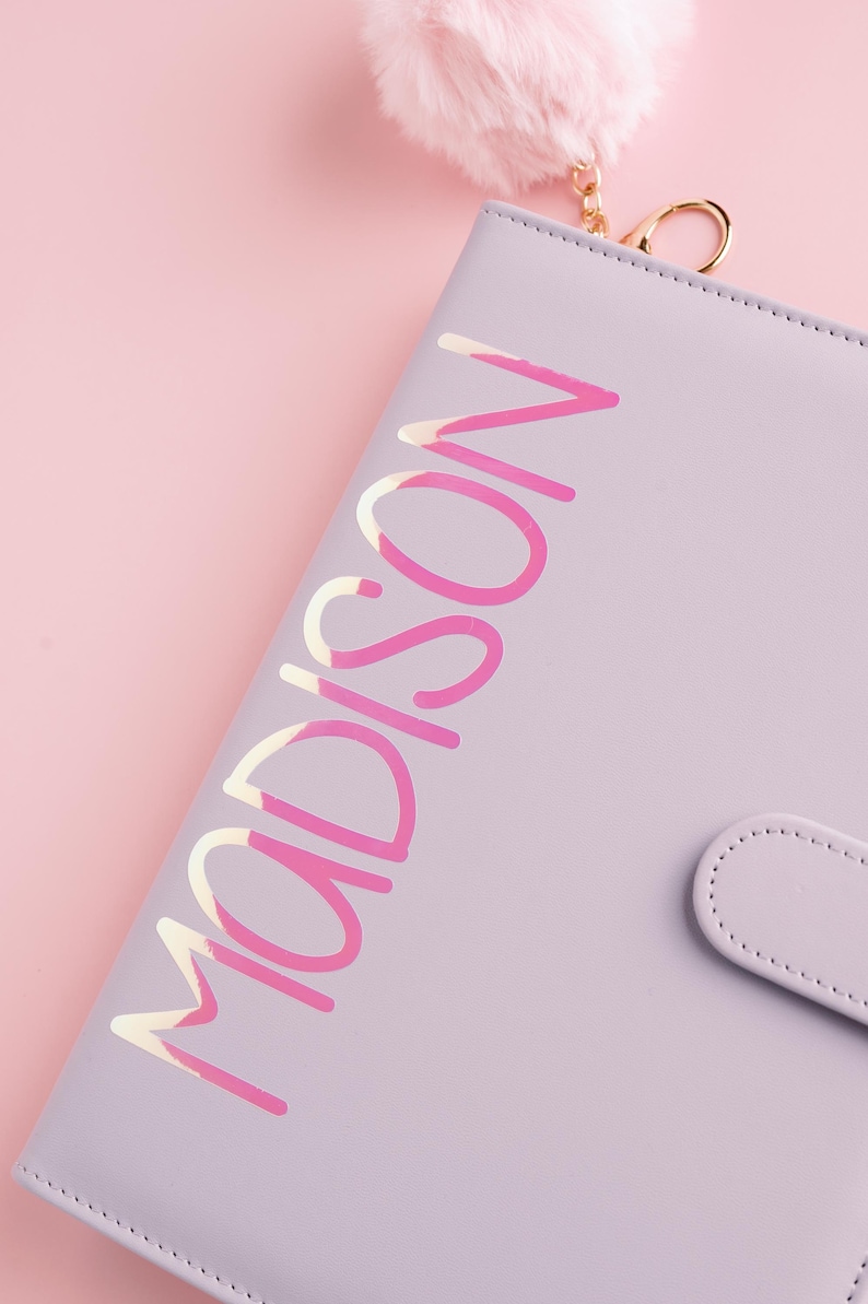May include: A lavender-colored planner with the name "MADISON" in a pink and white script. The planner has a matching snap closure and a fluffy pink keychain. The background is a soft pink.