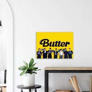 May include: A yellow canvas print featuring the word "Butter" in black script. Below the text are illustrations of seven people holding signs. The print is displayed on a white wall.