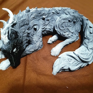 May include: A grey and black wolf figurine in a curled, resting position. The creature features intricate, swirling patterns across its fur, sharp spikes along its back, and prominent, branching horn-like structures extending from its head.