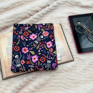 May include: A dark blue fabric book cover with a floral and skeleton design, resting on an open book. A black tablet with glasses is next to the book. The cover has a button closure.