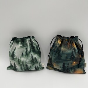 Peut inclure: Deux pochettes en tissu à cordon de serrage avec des motifs de forêt. La pochette de gauche présente un motif de pins verts dans la brume, tandis que celle de droite montre une forêt sombre avec des lumières dorées. Cordons noirs.