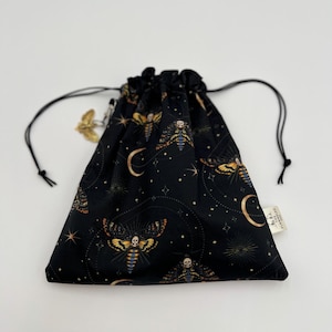 May include: Black drawstring bag with a repeating pattern of moths, moons, and stars. The moths have skull-like markings. The bag has a black drawstring closure and a small white tag. The bag is made of fabric.