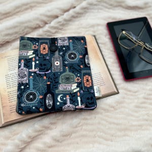 May include: A dark blue book cover with a spooky pattern featuring skulls, candles, and potion bottles, resting on an open book. A black tablet with glasses is next to the book. The items are on a fluffy, cream-coloured surface.