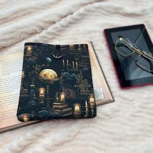 May include: A black and gold book cover with a celestial design featuring a globe, candles, and planets. The cover is open over a book, with a tablet and glasses in the background. The scene is set on a soft, white, textured surface.