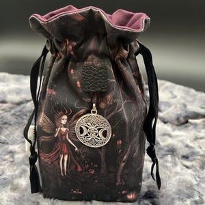Peut inclure: Une pochette à cordon avec un imprimé de forêt sombre, une illustration de fée et un pendentif en métal argenté représentant une triple lune et un pentacle. Le sac possède une doublure intérieure violet foncé et des cordons noirs.