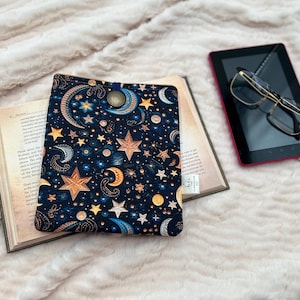 May include: A navy blue book sleeve with a celestial design featuring gold stars, moons, and paisley accents. The sleeve rests on an open book, with a tablet and glasses nearby. The background is a soft, textured, light-coloured fabric.