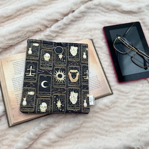 May include: A black and gold tarot card print book cover with a button closure, on an open book. A black tablet with glasses is beside the book. The background is a soft, textured, light-coloured fabric.
