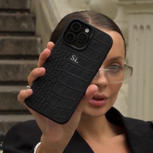 May include: Black phone case with a crocodile skin effect. The case features a camera aperture and the initials "SL". The case is held up in front of a person.